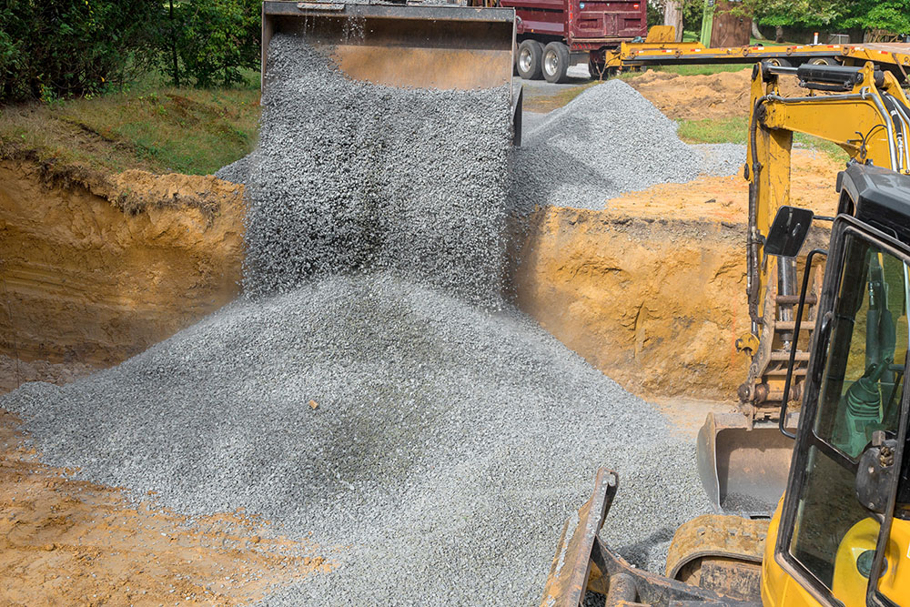 Excavator fills irregularities in excavations with granite rubble base that is then covered by concrete foundations in construction site